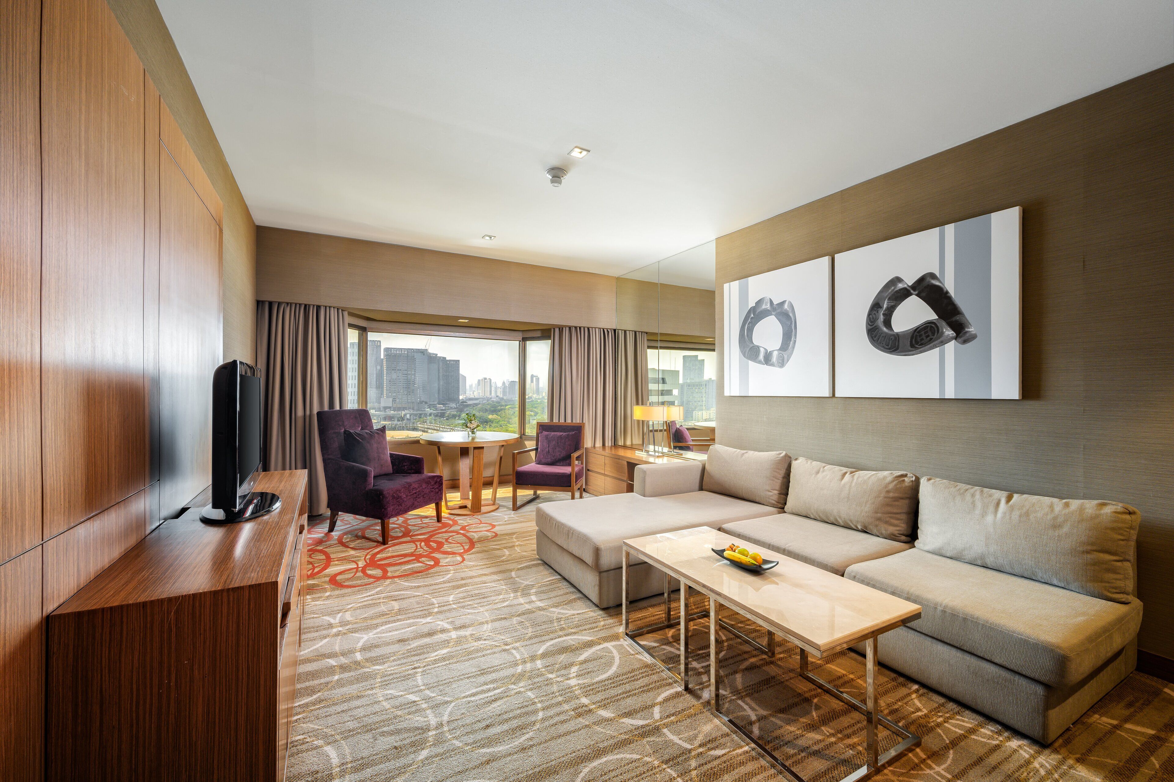 A modern hotel room with a large sofa, a coffee table, a TV, and a city view through the window.