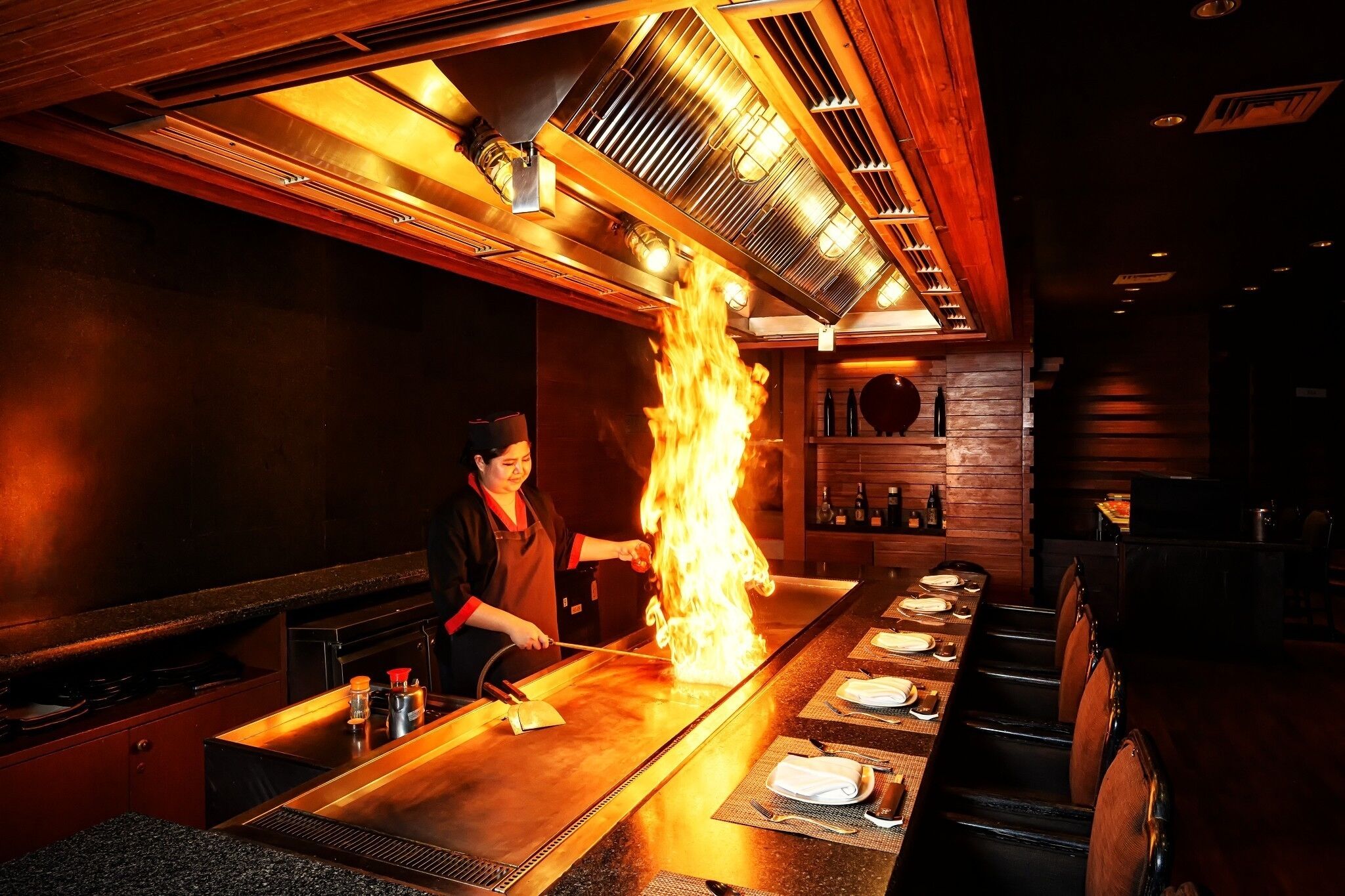 Un chef realizando una técnica de cocina a la parrilla en un restaurante con una barra larga y un techo alto.