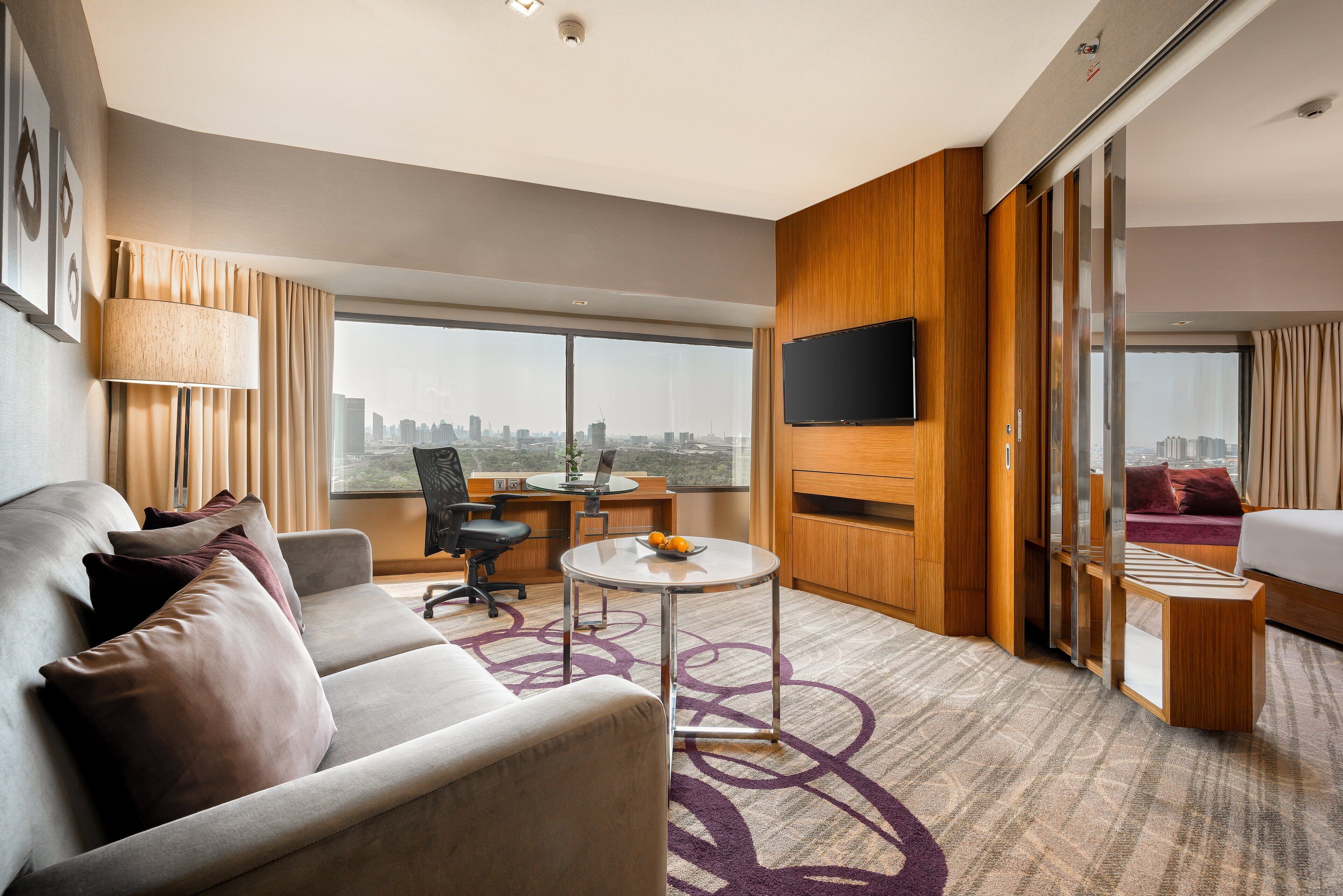 A modern hotel room with a sofa, a desk, a flat-screen TV, and a city view.