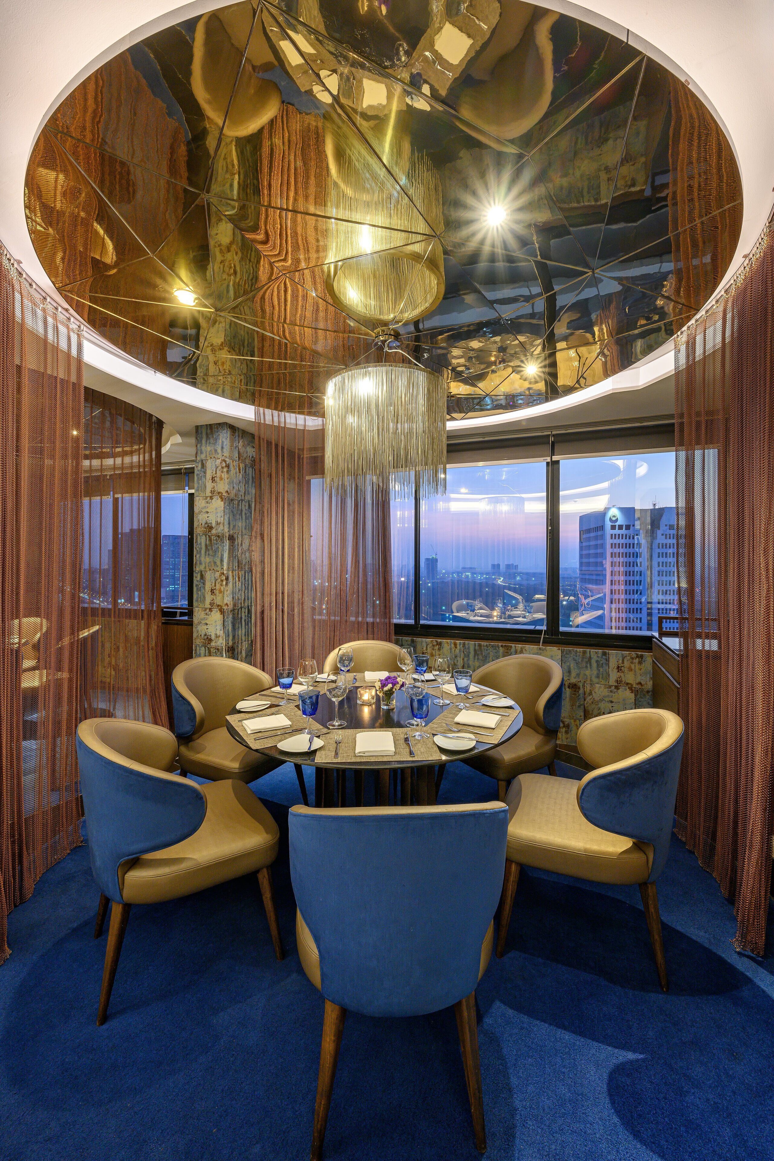 A round table with six chairs, a chandelier, and a city view through large windows.
