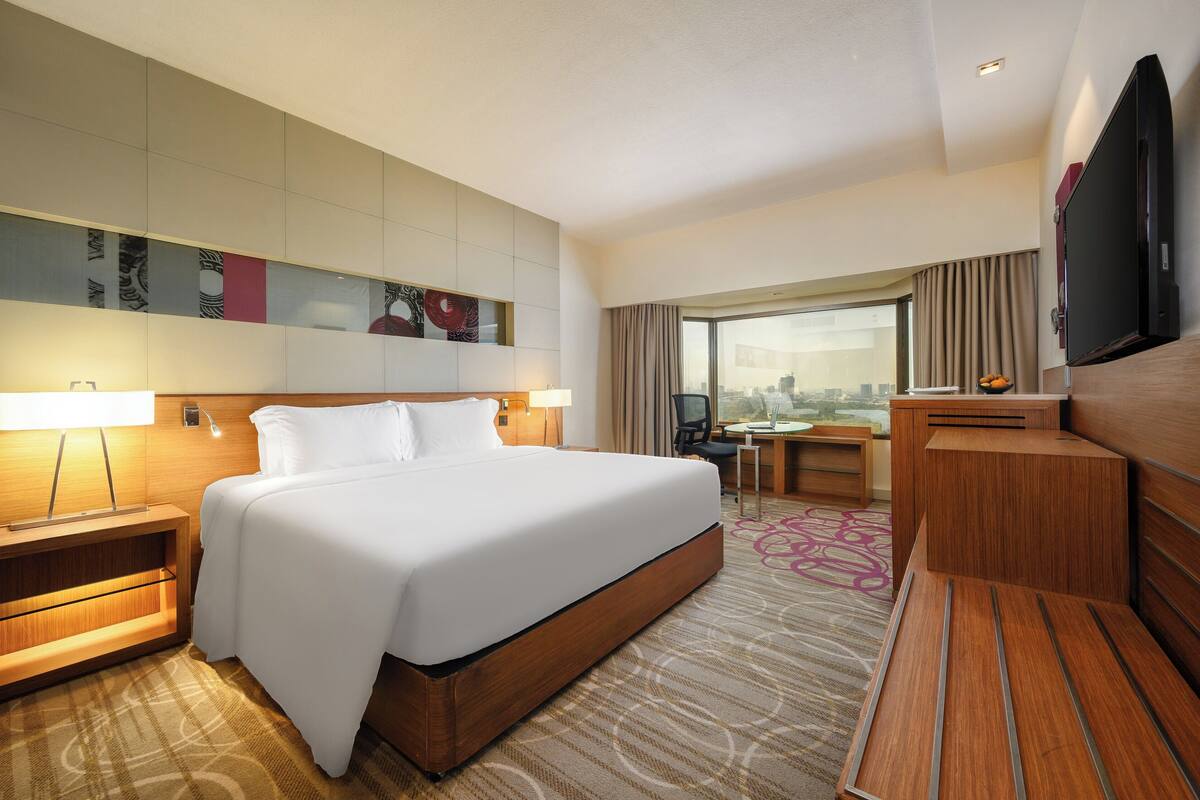 Ein Hotelzimmer mit einem großen Bett, einem Schreibtisch, einem Sessel, einem Fernseher und Blick auf die Stadt.