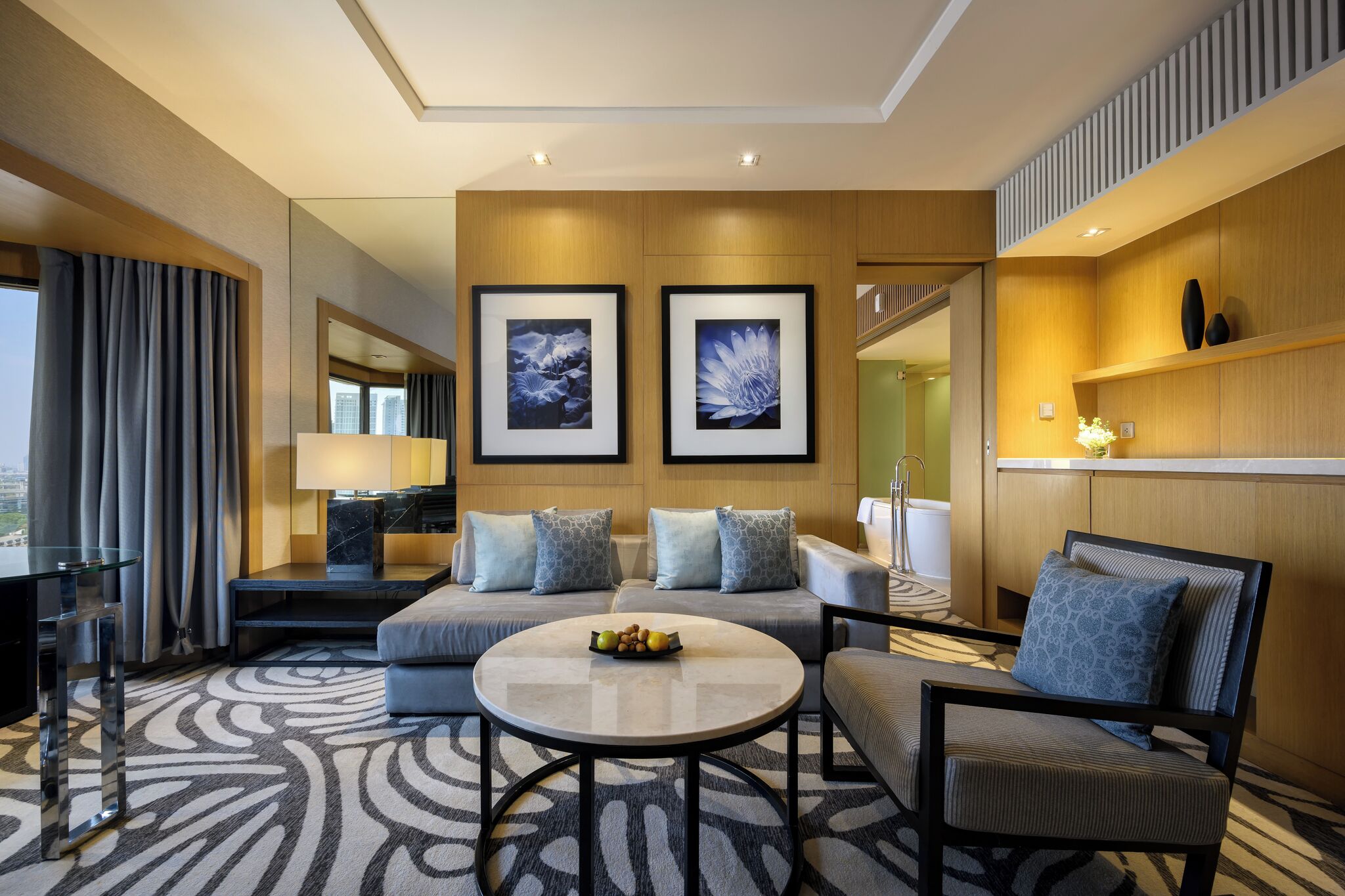 Une chambre d’hôtel moderne avec un canapé, une table basse ronde sur laquelle sont disposés des fruits, un téléviseur et des œuvres d’art accrochées au mur.