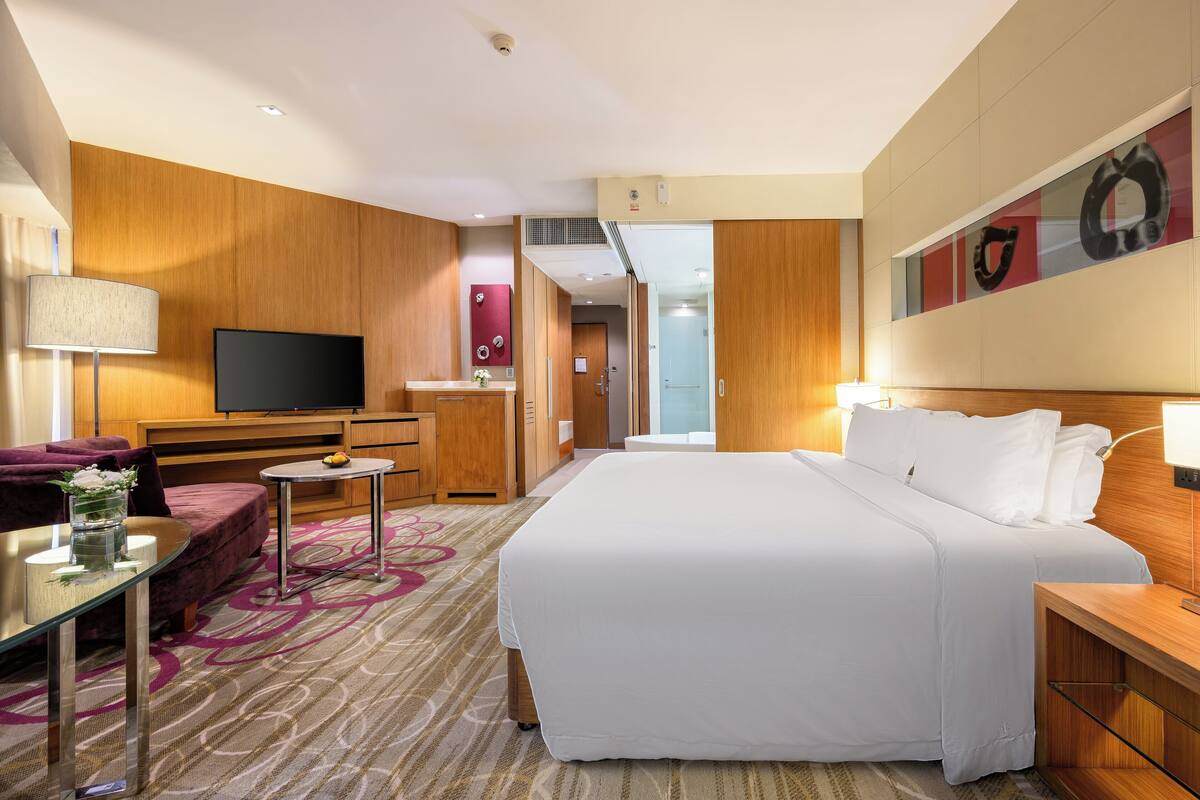 Ein Hotelzimmer mit einem großen Bett, einem Fernseher, einem Schreibtisch, einer Sitzecke und einem durchgegangenes Badezimmereingang sichtbaren Badezimmer.