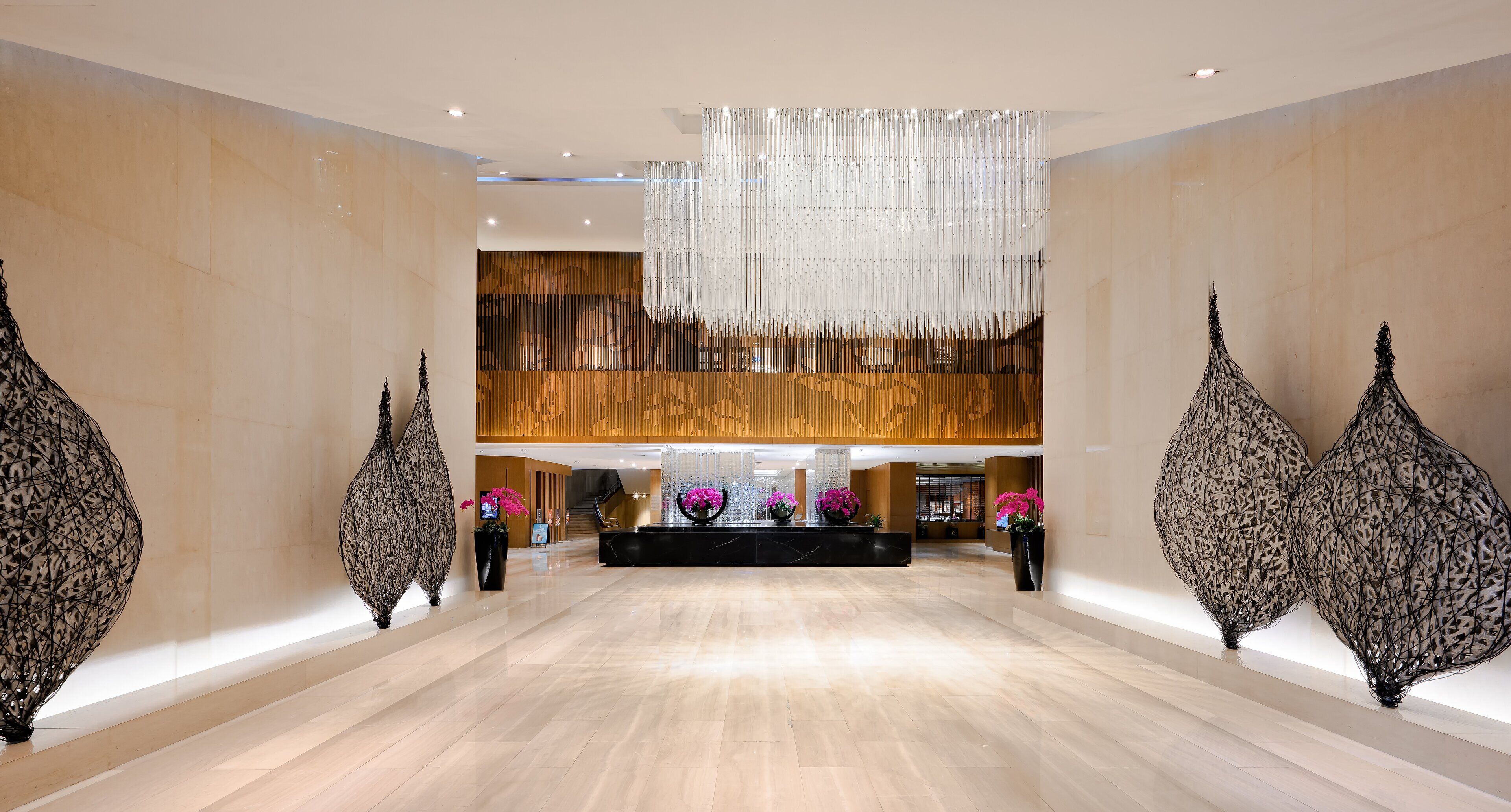 A spacious lobby with a central reception desk, large decorative wall panels, and a modern chandelier.