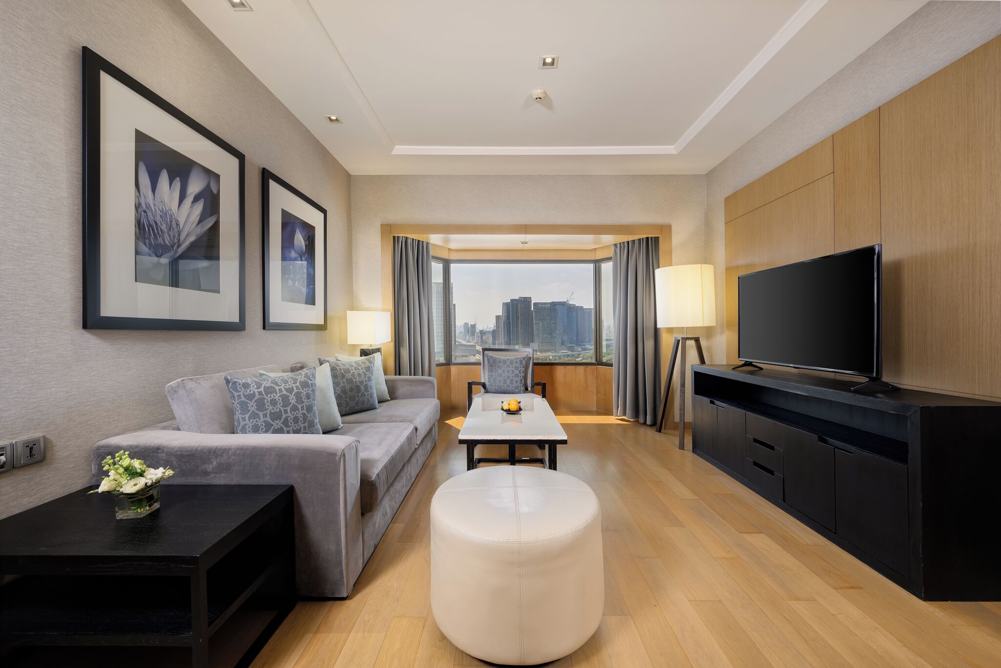 A modern hotel room with a sofa, a round ottoman, a TV on a cabinet, and framed artwork on the wall.