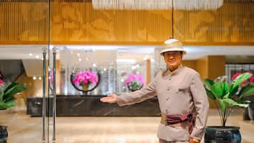 A person in a beige suit and hat standing in a modern lobby with a glass partition, large potted plants, and a decorative ceiling.