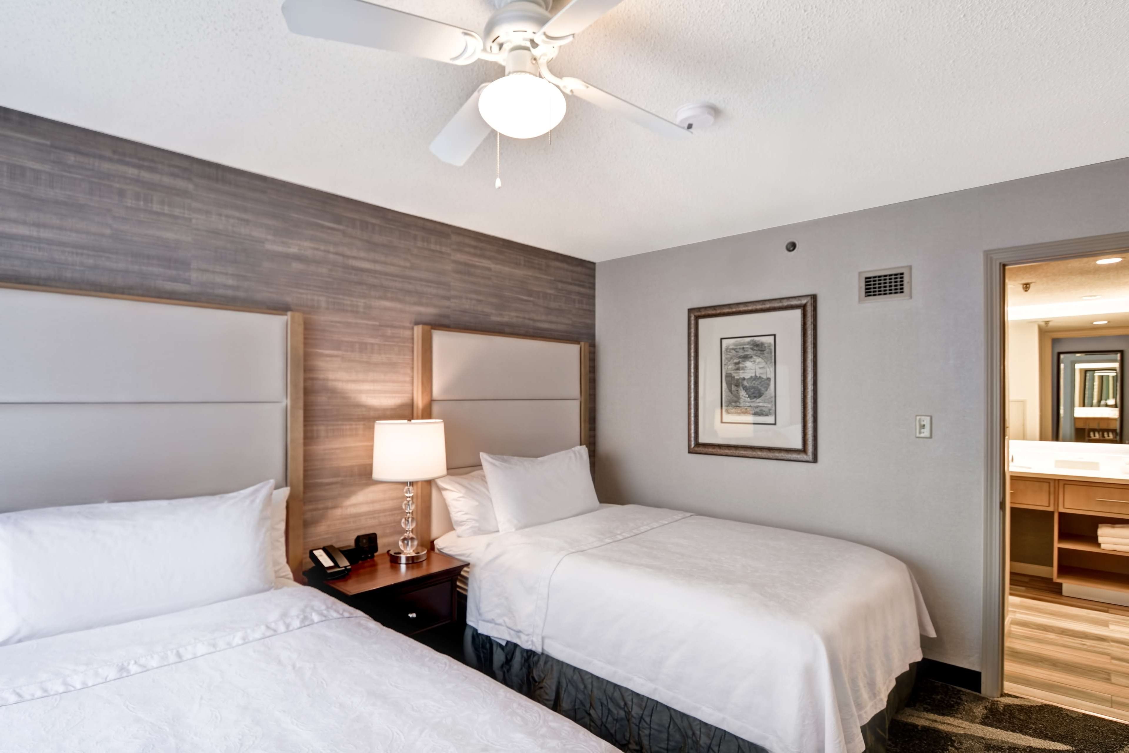 A hotel room with two beds, a ceiling fan, a nightstand, a lamp, and a framed picture on the wall.