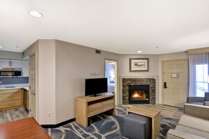 Una sala de estar moderna con chimenea, un televisor sobre un soporte de madera, una zona de cocina con microondas y una puerta que da a otra habitación.