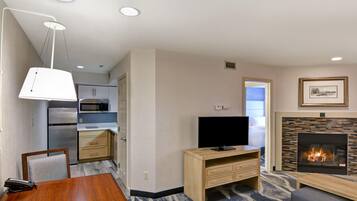 A modern hotel room with a kitchenette, a flat-screen TV, a fireplace, and a dining area.