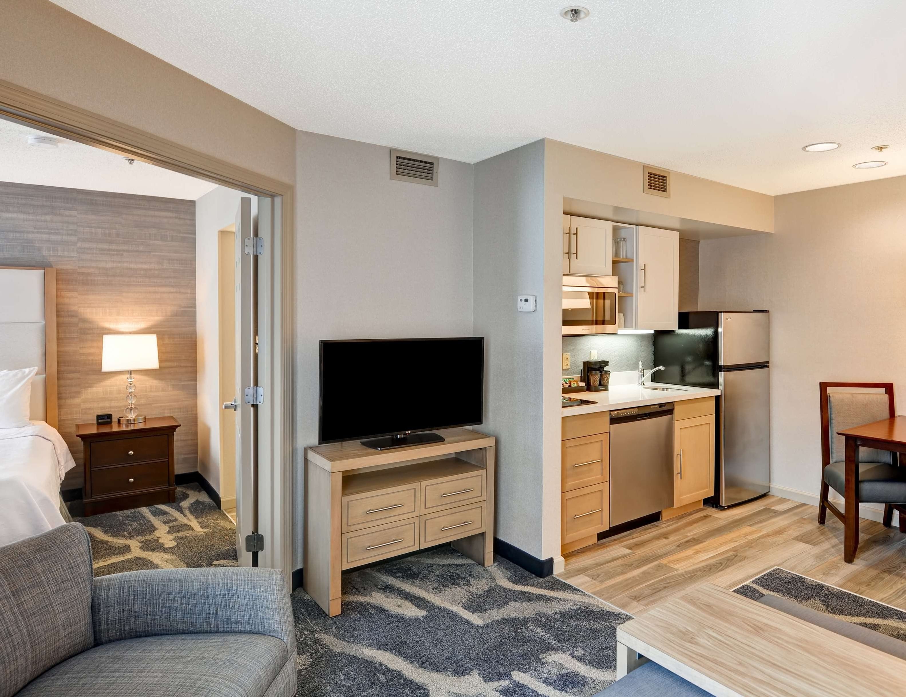 A hotel room with a bed, a television on a wooden stand, a kitchenette with a refrigerator, microwave, and sink, and a small dining table with a chair.