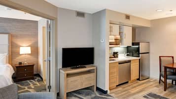 A hotel room with a bed, a television on a wooden stand, a kitchenette with a refrigerator, microwave, and sink, and a small dining table with a chair.