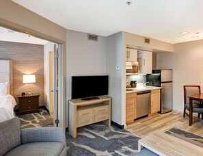 Une chambre d’hôtel comprenant un lit, un téléviseur posé sur un meuble en bois, une kitchenette équipée d’un réfrigérateur, d’un four à micro-ondes et d’un évier, ainsi qu’une petite table à manger avec une chaise.