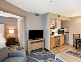 Habitación de hotel con cama, televisor sobre un soporte de madera, kitchenette con refrigerador, microondas y lavaplatos, y una pequeña mesa de comedor con una silla.