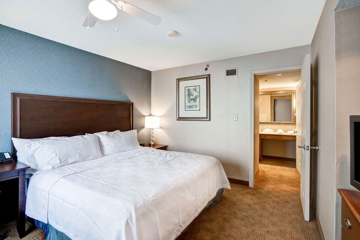A hotel room with a large bed, a nightstand, a bedside lamp, a framed picture on the wall, and an open door to the bathroom.