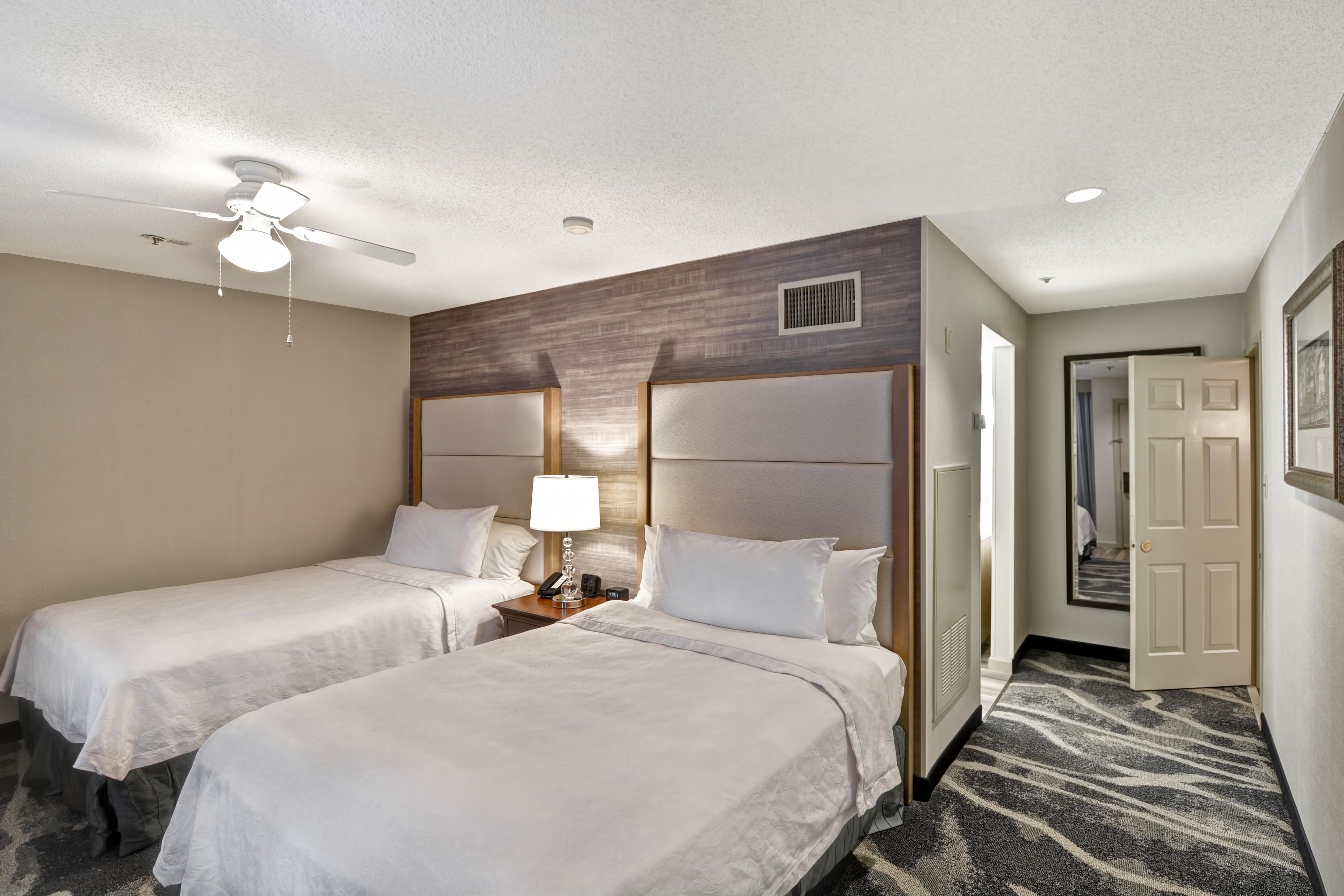 A hotel room with two beds, a ceiling fan, a nightstand with a lamp, and a door leading to another room.