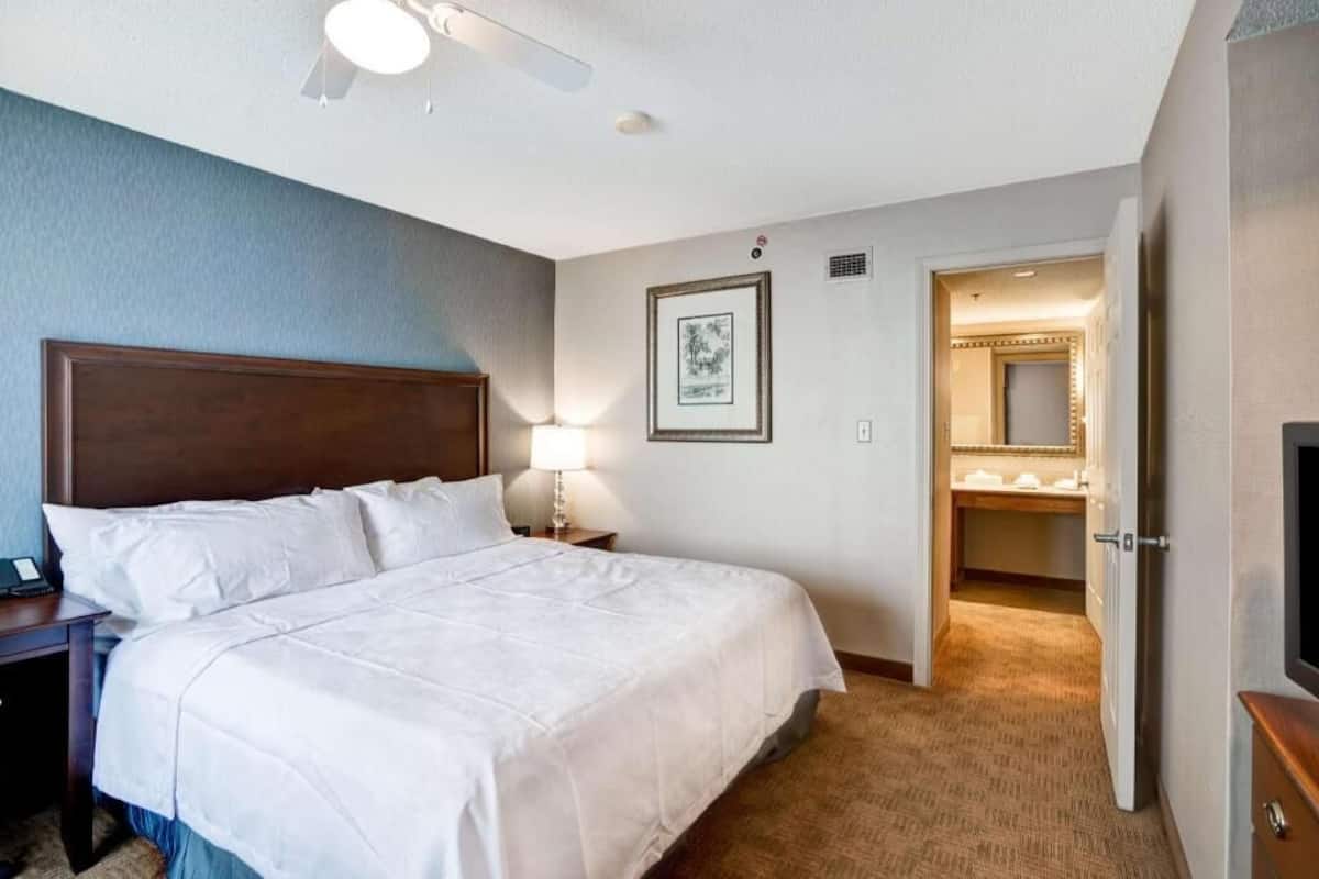 Habitación de hotel con una cama grande, una mesita de noche, una lámpara, un cuadro en la pared y un baño visible a través de una puerta abierta.