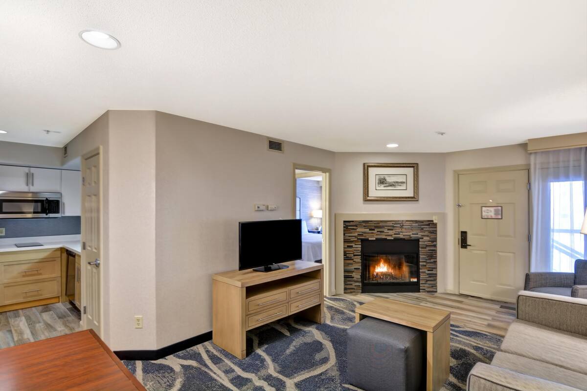 Una sala de estar con chimenea, un televisor sobre un mueble de madera, una mesa de comedor y una zona de cocina con armarios y un microondas.