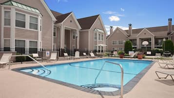 A swimming pool with lounge chairs and a fence, surrounded by multi-story buildings with large windows and a clear sky.
