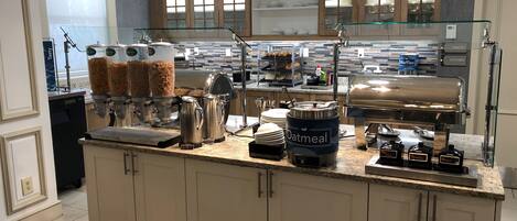 A breakfast bar with coffee machines, a waffle maker, and various food containers.