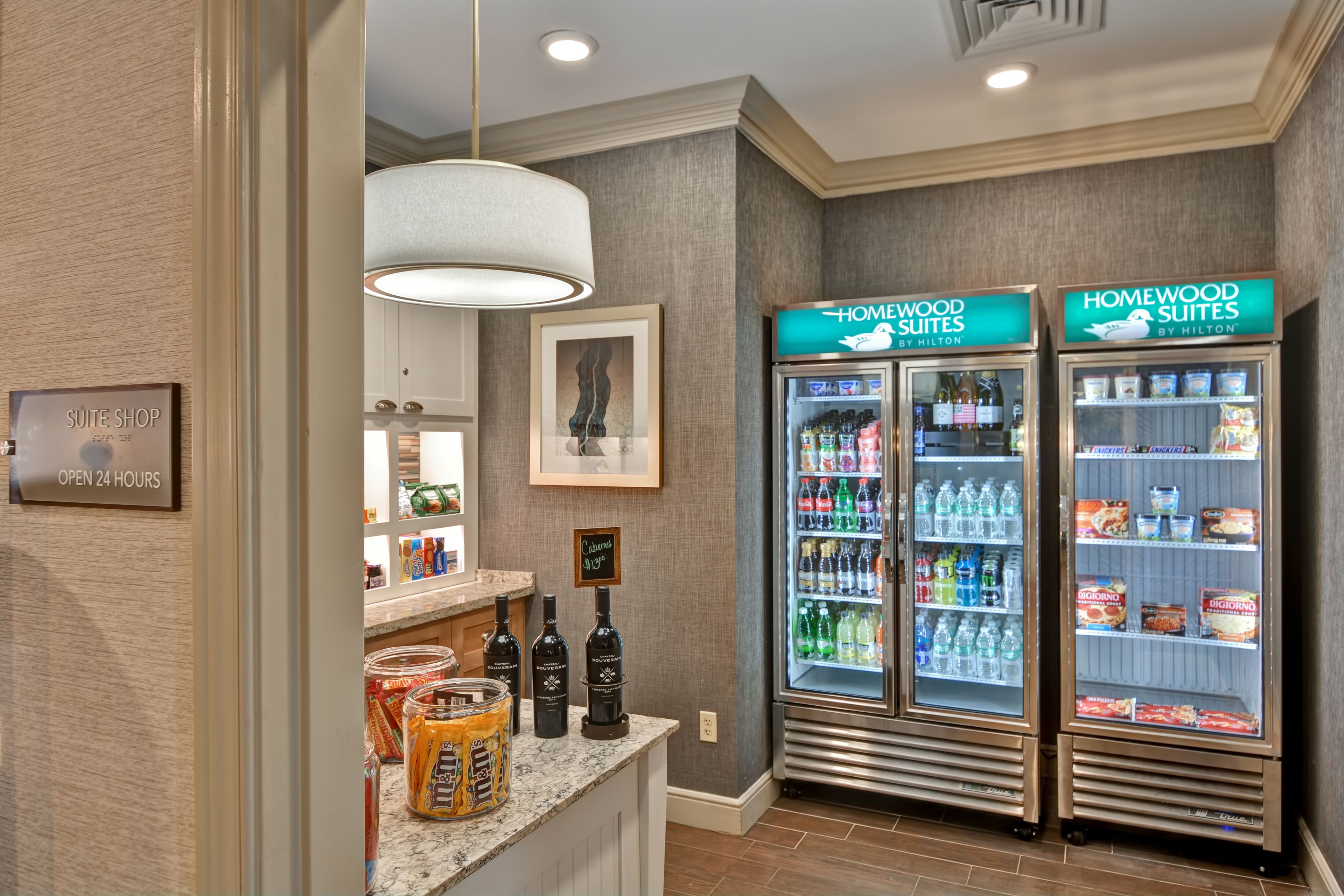 A hotel suite with a minibar, a sign for the suite shop, and two Homewood Suites refrigerators stocked with beverages and snacks.