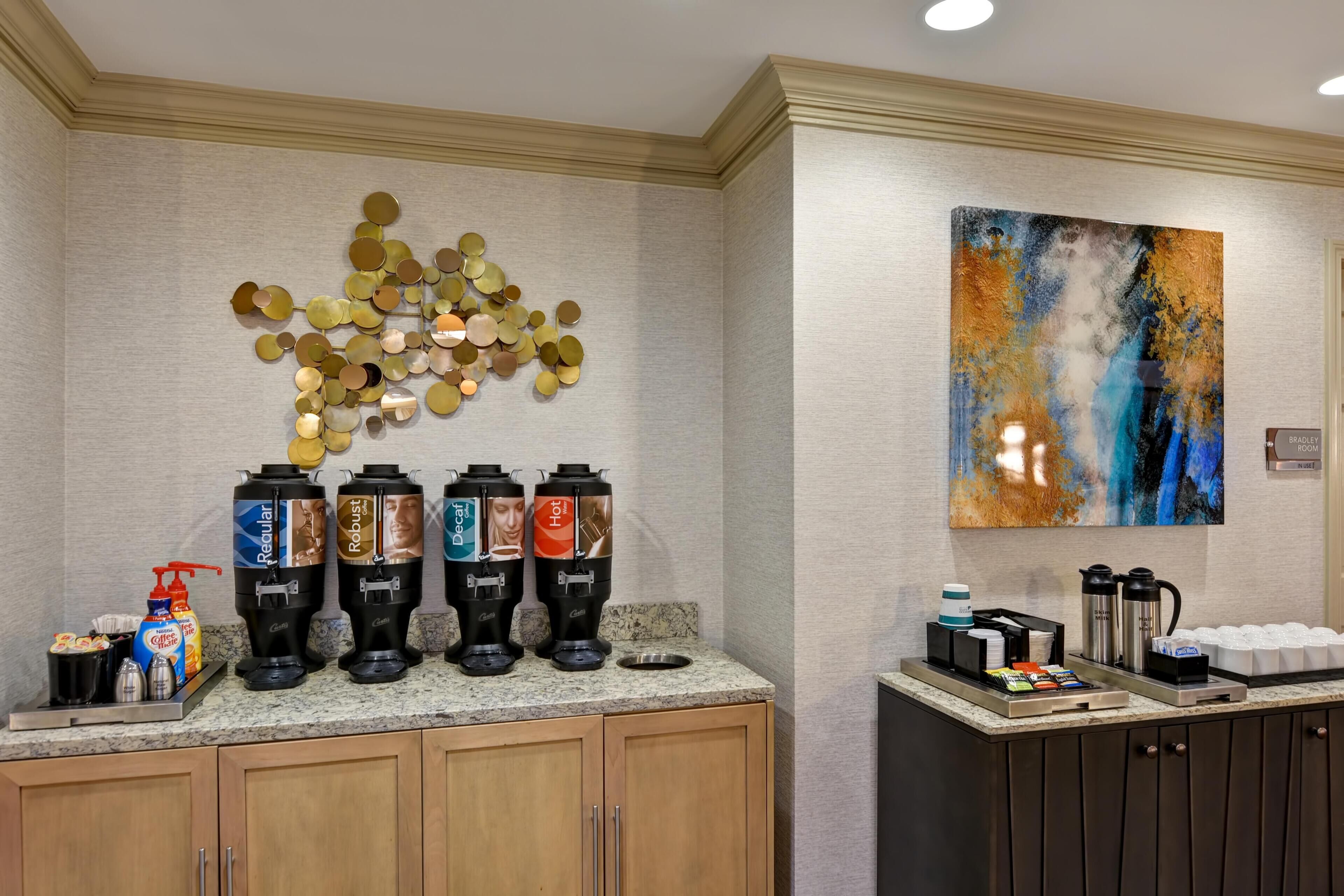 A coffee station with multiple dispensers, a painting, and a tray with coffee cups and a coffee pot.