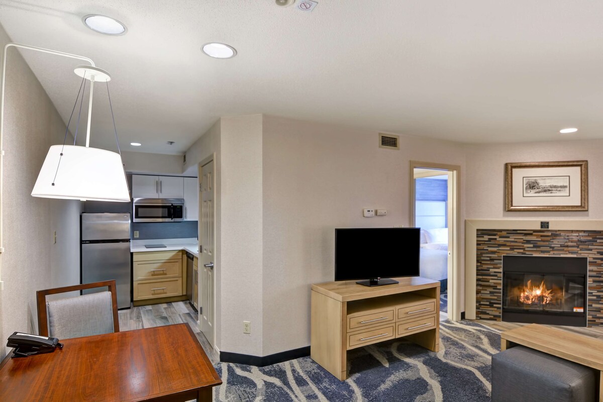 A hotel room with a dining table, a TV, a fireplace, a kitchen area, and a patterned carpet.