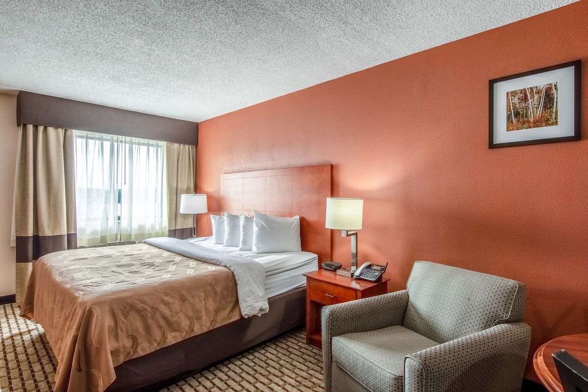 Une chambre d’hôtel avec un lit, deux lampes de chevet, une chaise, un bureau avec un téléphone et un tableau encadré au mur.