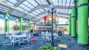 A spacious indoor area with a high ceiling and skylights, featuring multiple seating areas with tables and chairs, surrounded by green plants and a patterned carpet. - Country Inn & Suites by Radisson, Muskegon, MI (Muskegon)