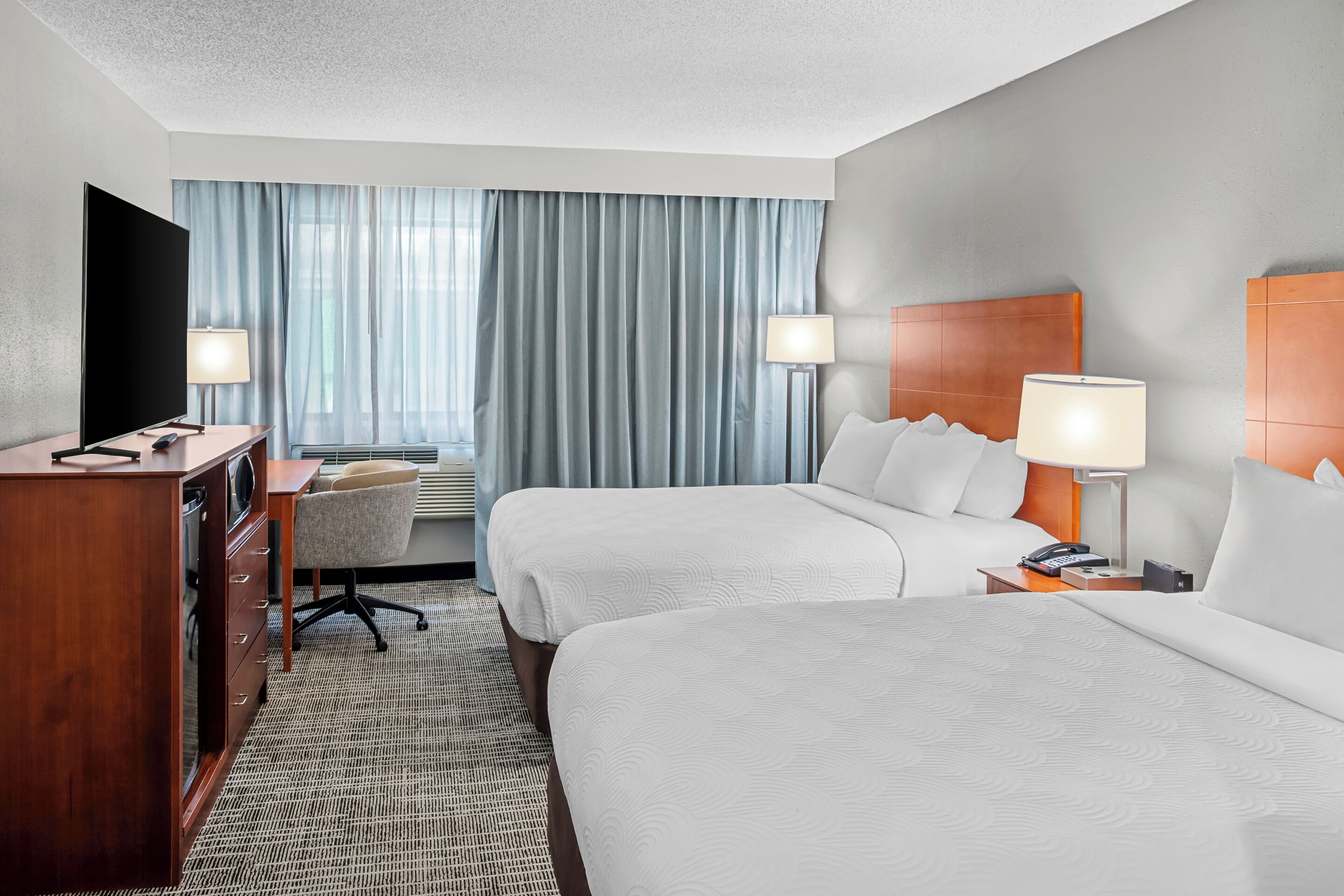 A hotel room with two beds, a desk, a TV, and a lamp.
