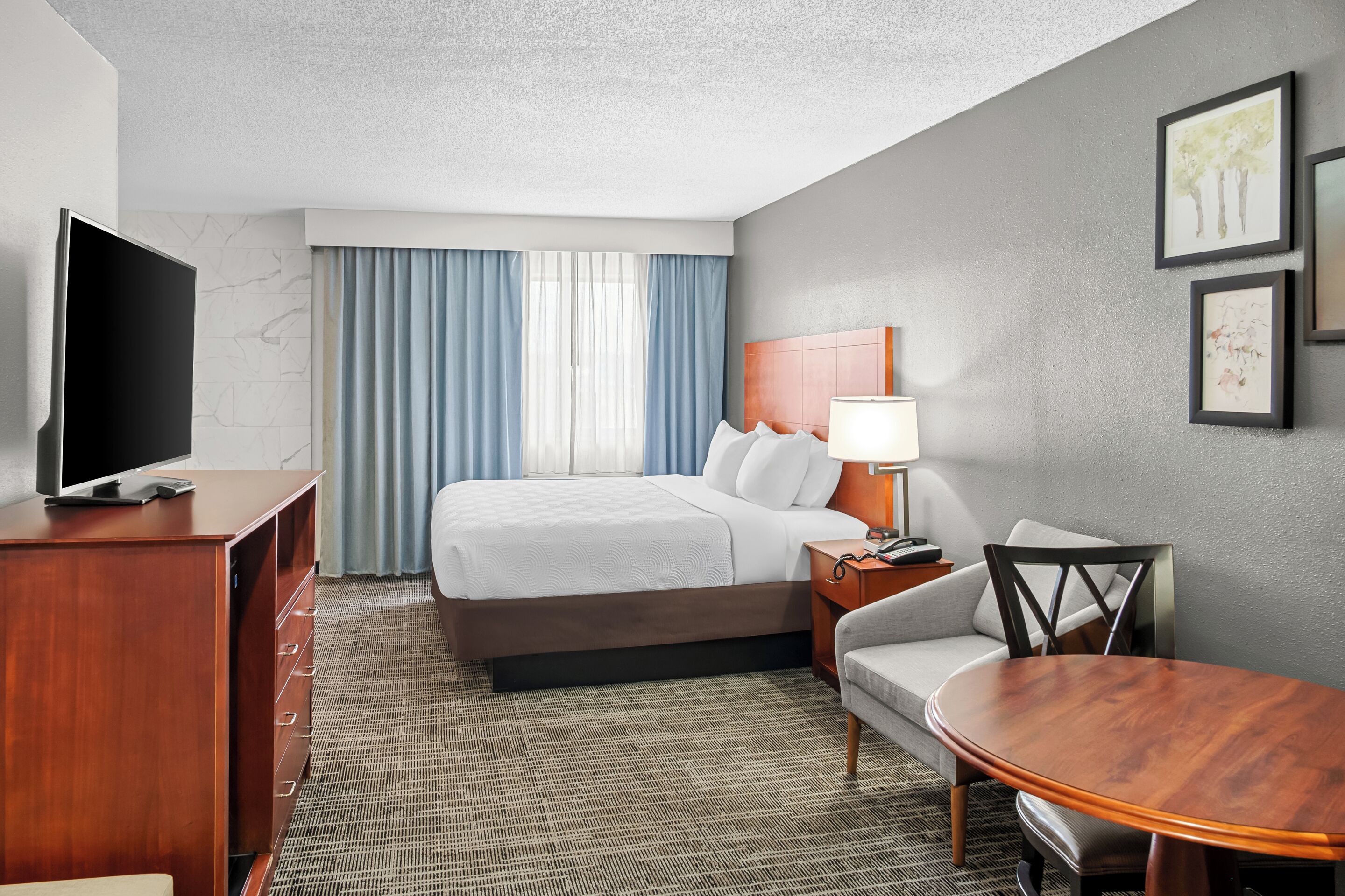 A hotel room with a bed, a TV, a desk with a lamp, a chair, and a small table.