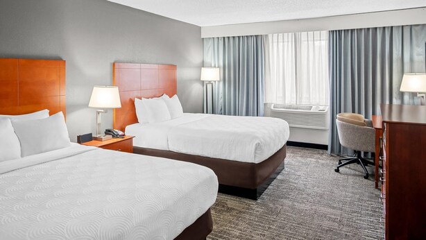 A hotel room with two beds, a desk, a chair, a lamp, and a window with curtains.