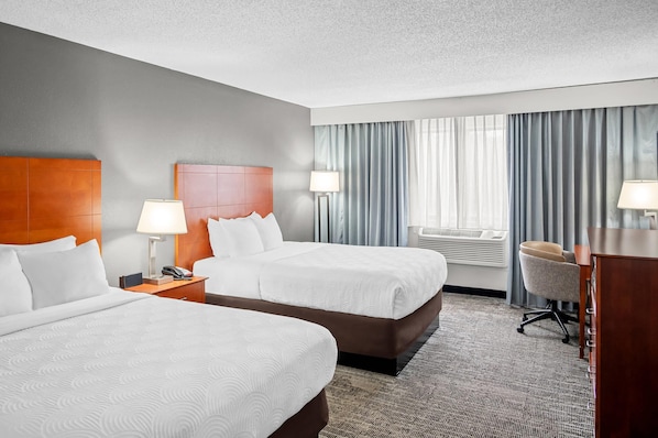 A hotel room with two beds, a desk, a chair, a lamp, and a window with curtains.