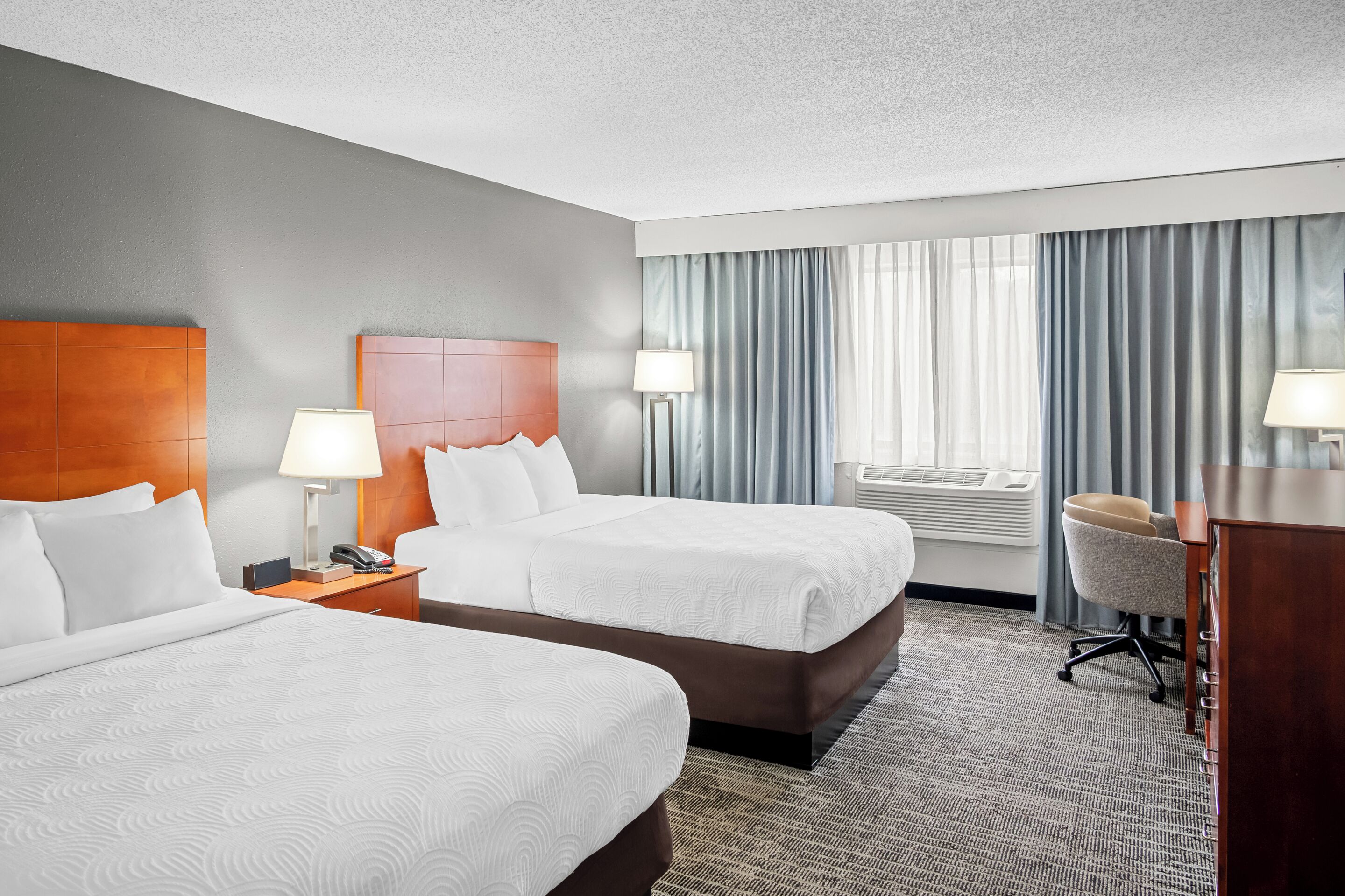 A hotel room with two beds, a desk, a chair, a lamp, and a window with curtains.