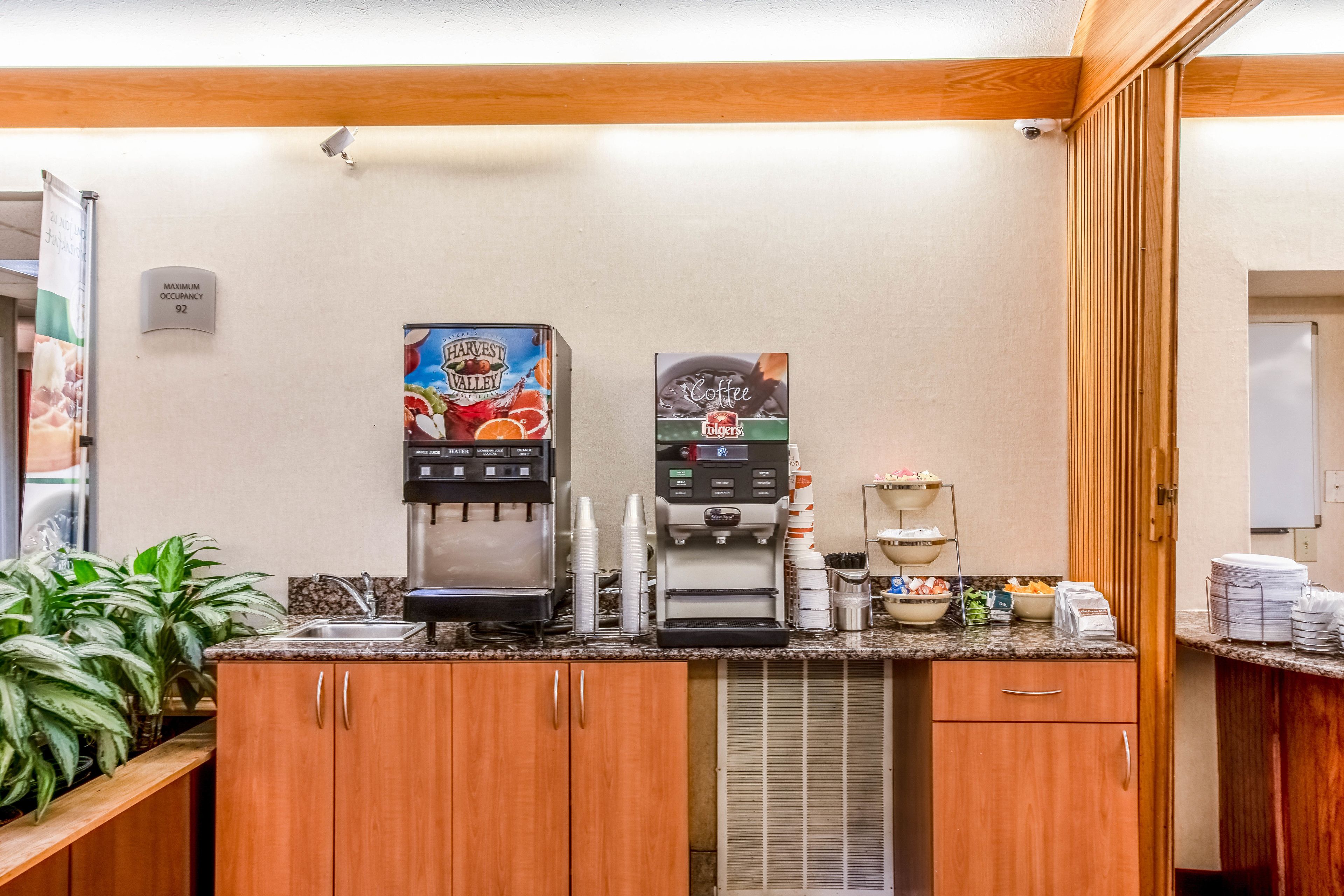 A kitchenette with a coffee machine, a water dispenser, and a fruit juice dispenser. There are cabinets, a sink, and a plant.