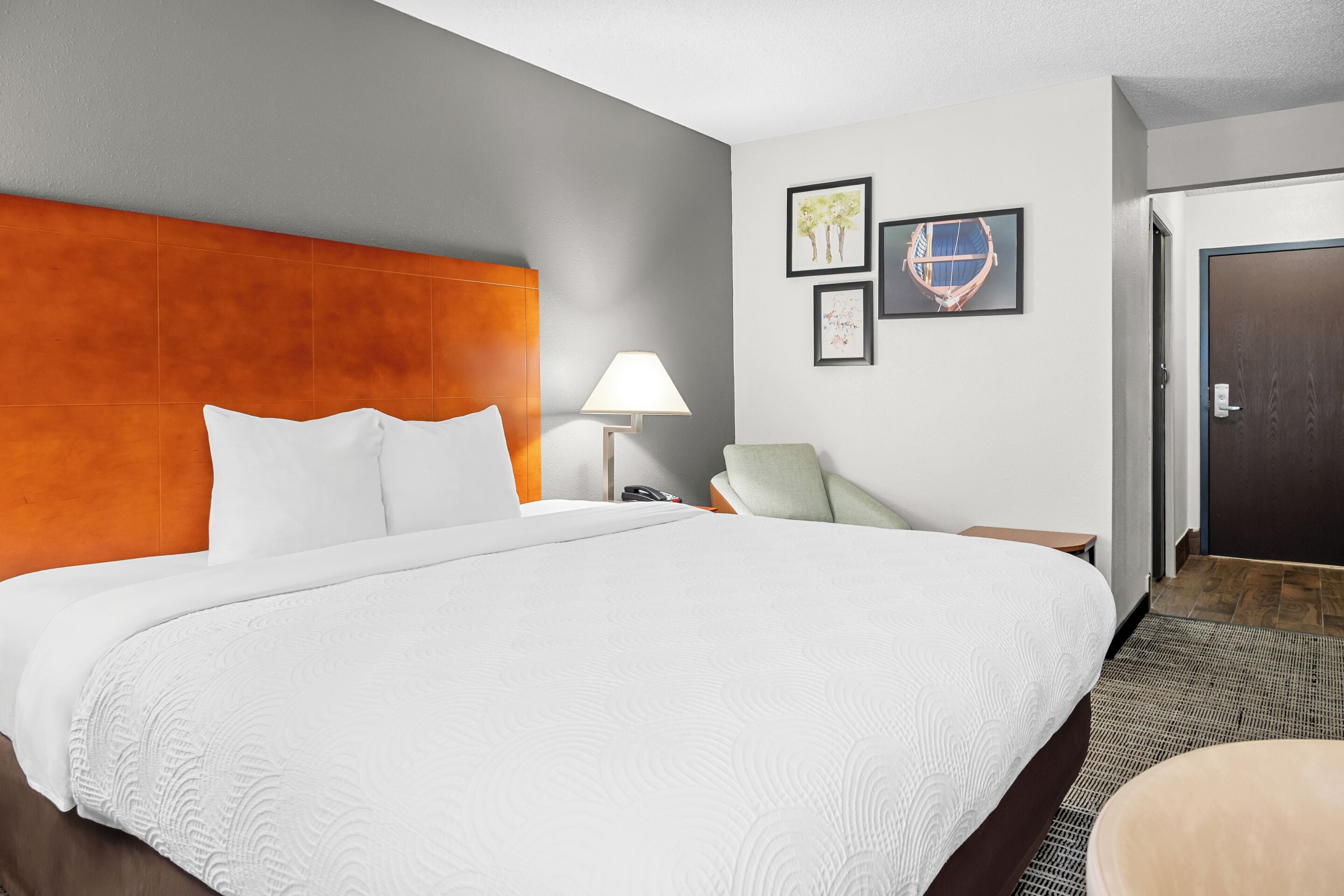 Habitación de hotel con una cama grande, una mesita de noche con lámpara, una silla y una puerta.