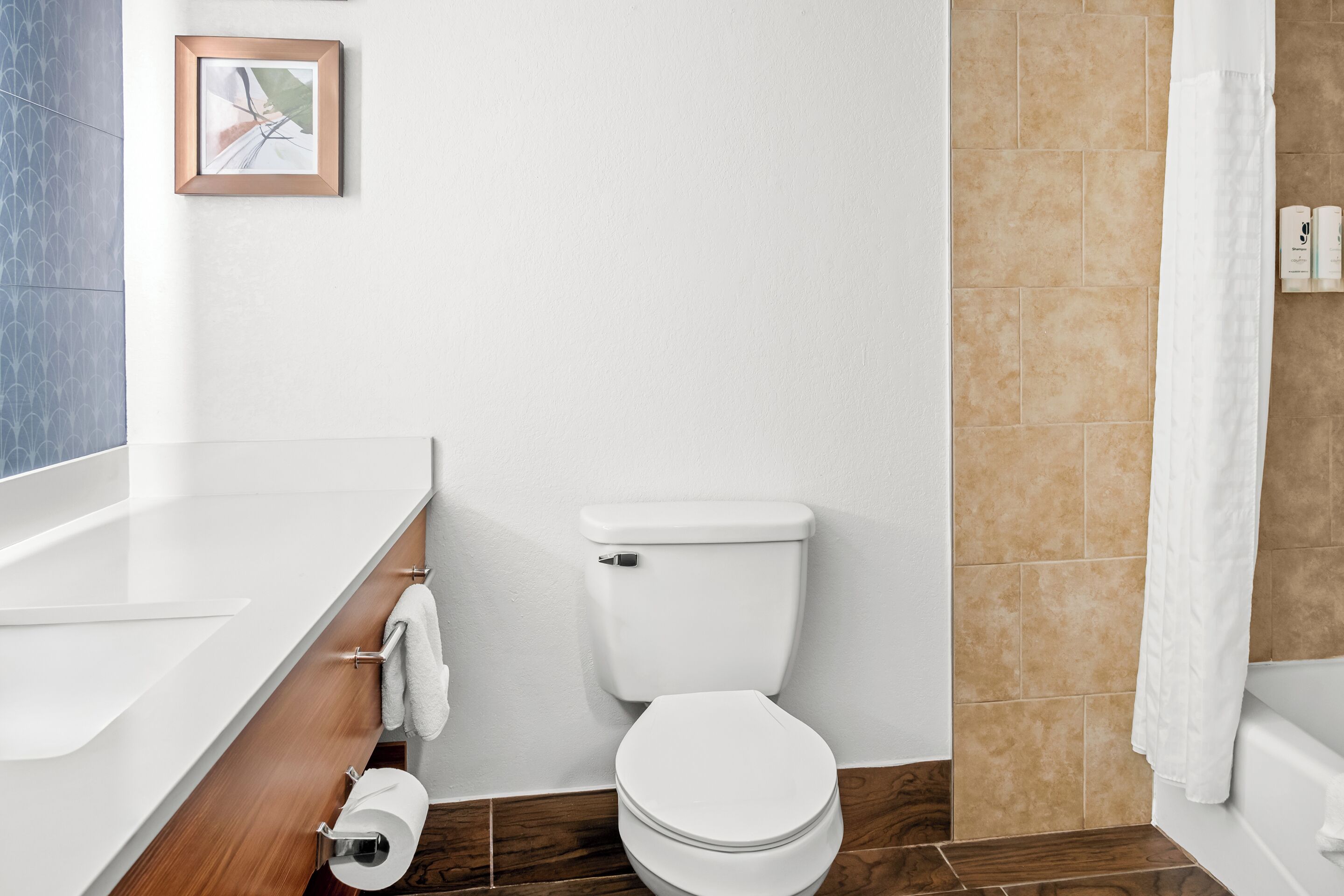 Et moderne badeværelse med et hvidt toilet, en trækommode, et billede i ramme på væggen og en brusevæg.