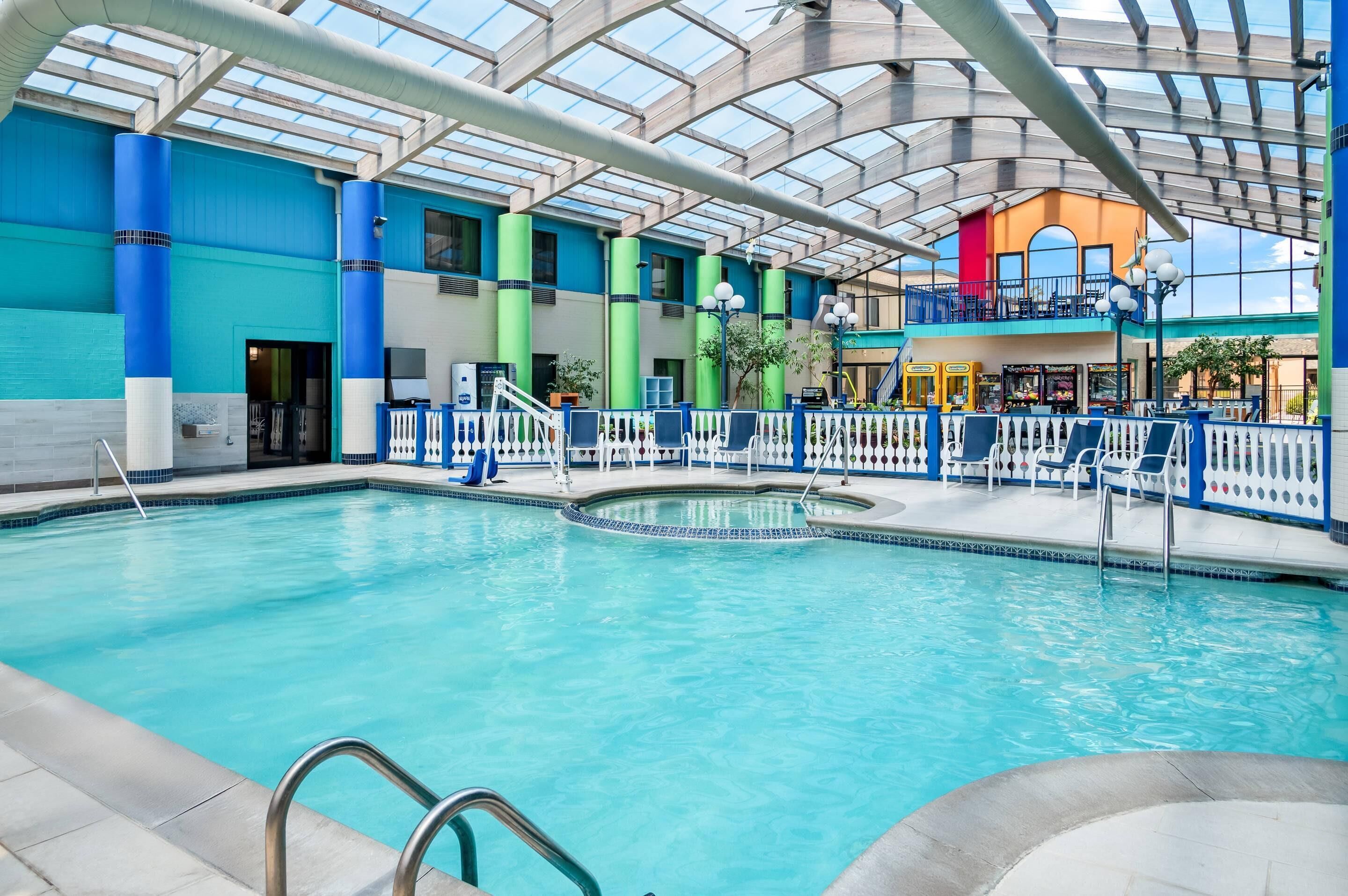 An indoor swimming pool with a glass ceiling, blue and green pillars, and a fenced area with lounge chairs.