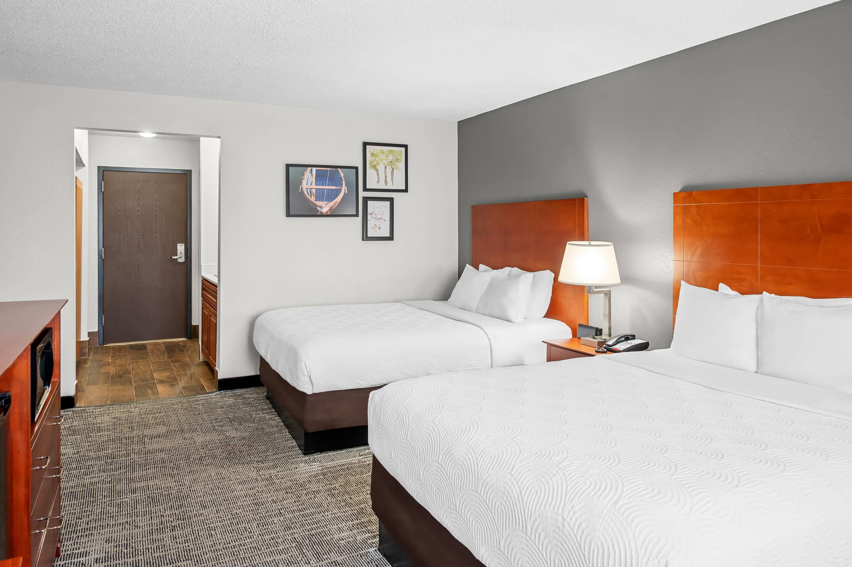 A hotel room with two beds, a wooden headboard, a bedside table with a lamp, and a door leading to a hallway.
