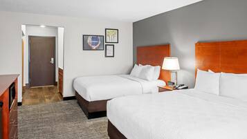 A hotel room with two beds, a wooden headboard, a bedside table with a lamp, and a door leading to a hallway.