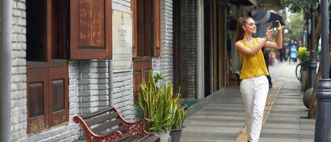 A woman taking a selfie on a sidewalk next to a building with a brick facade, a bench, and potted plants.