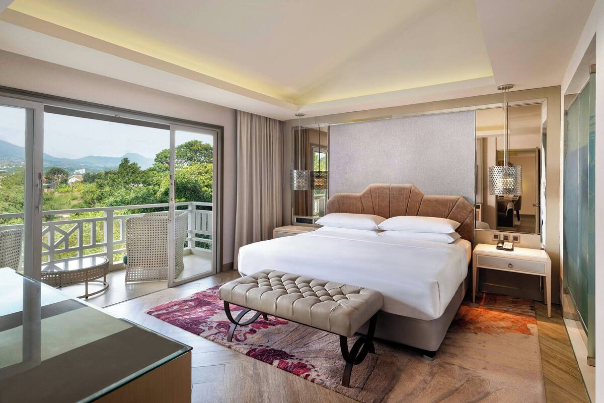 Une chambre d’hôtel moderne dotée d’un grand lit, d’une table de chevet, d’un bureau et d’un balcon offrant une vue sur la verdure et les montagnes.