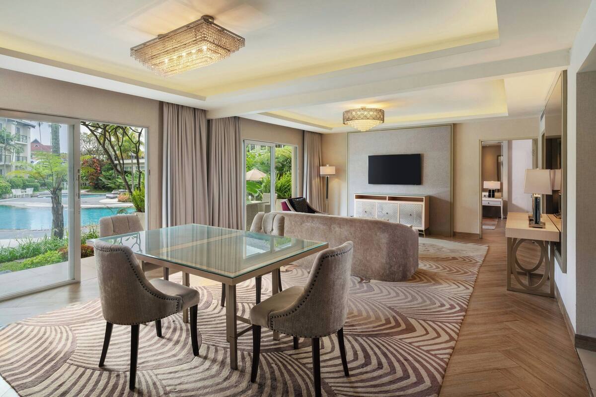 Une chambre d’hôtel moderne avec une table à manger en verre, deux fauteuils rembourrés, un canapé, un téléviseur à écran plat et une vue sur une piscine et un espace verdoyant.