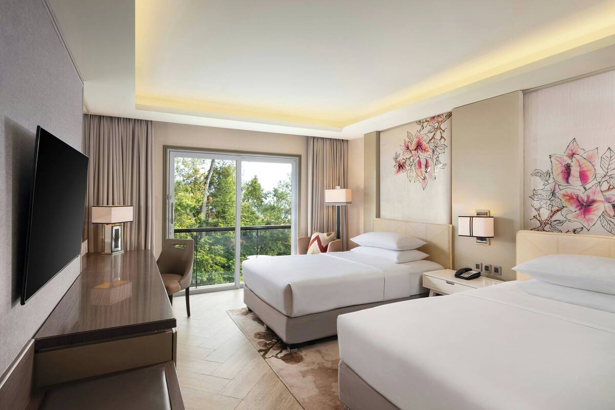 Une chambre d’hôtel avec un grand lit, un bureau doté d’un téléviseur à écran plat, une chaise et un balcon offrant une vue sur les arbres.