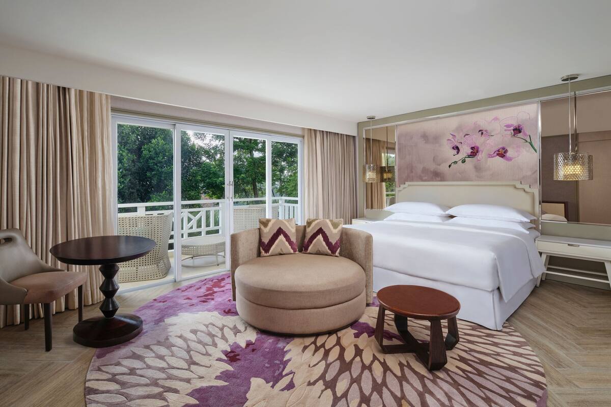 Une chambre d’hôtel avec un grand lit, un fauteuil rond, une petite table et un balcon agrémenté d’un fauteuil en osier.