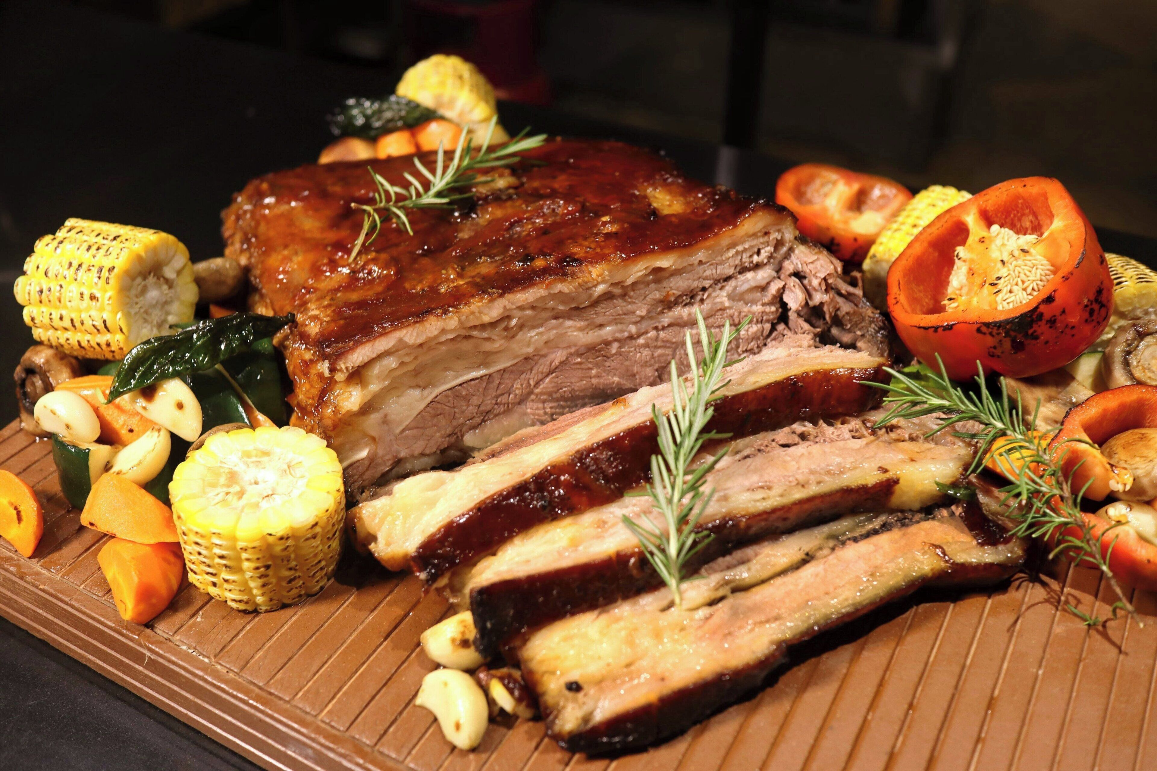A roasted pork loin with ribs, garnished with rosemary, served with vegetables like corn, carrots, and squash.