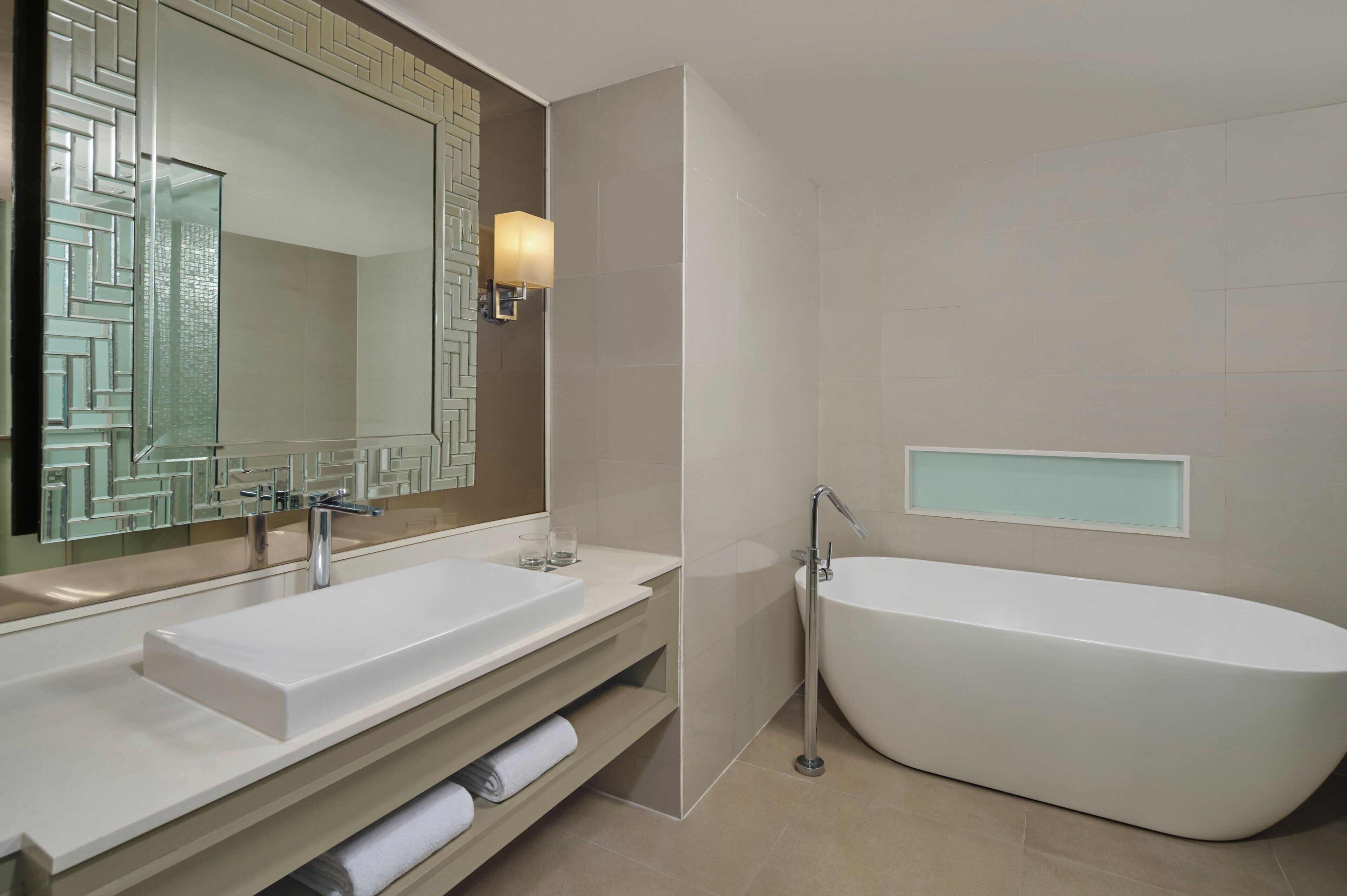 Un baño moderno con una bañera independiente, un espejo grande con un revestimiento geométrico y un mueble de tocador con toallero.