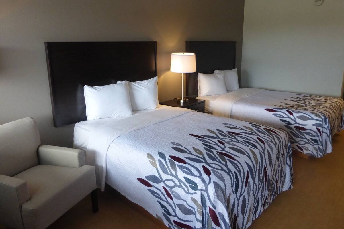 A hotel room with two beds, a nightstand with a lamp, a chair, and a headboard.