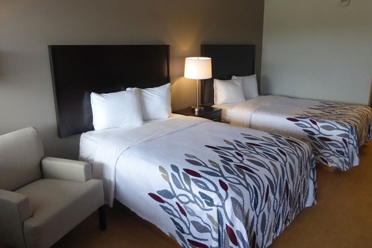A hotel room with two beds, a nightstand, a lamp, and a sofa.
