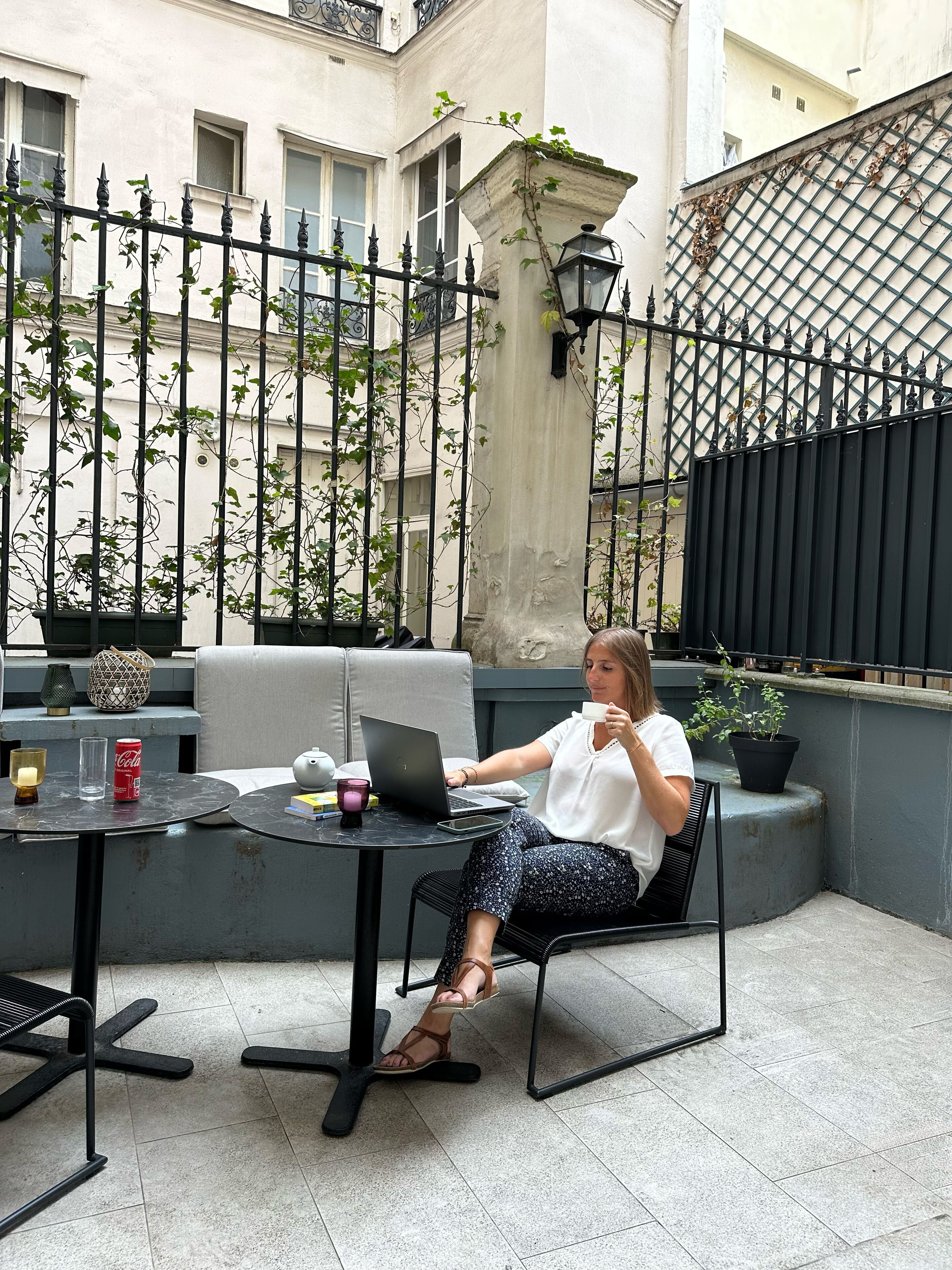 A woman sitting at a table with a laptop, a cup, and a Coca-Cola can. The table is on a balcony with a view of a building and a fence.