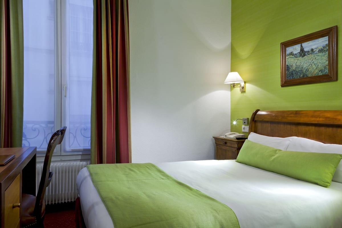A hotel room with a bed, a desk, a chair, a nightstand, a lamp, and a painting on the wall.